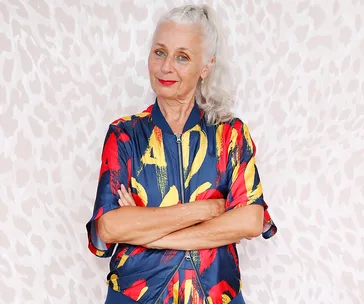 Elderly woman in a colorful jacket, arms crossed, standing against a patterned background.