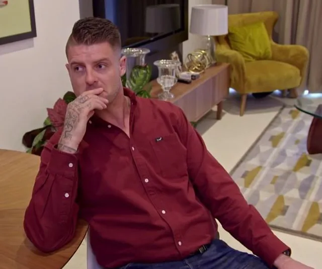 Man in a red shirt sitting thoughtfully in a modern living room with a hand near his face.