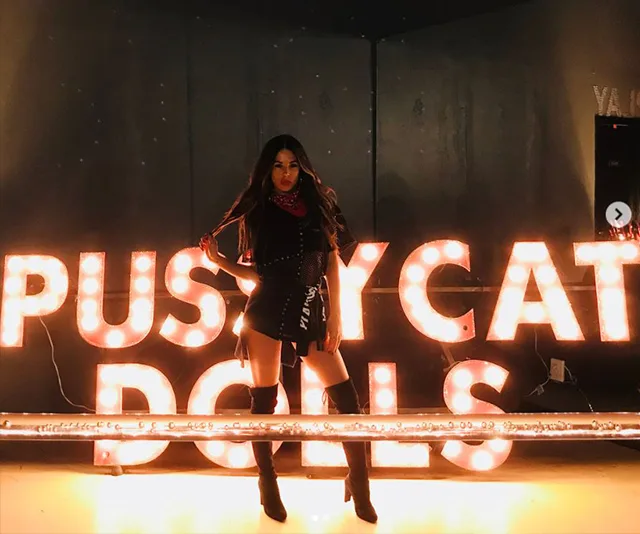 Woman poses confidently in front of the glowing "Pussycat Dolls" sign, wearing a stylish outfit and high boots.