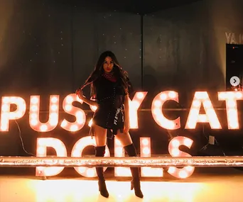 Woman poses confidently in front of the glowing "Pussycat Dolls" sign, wearing a stylish outfit and high boots.
