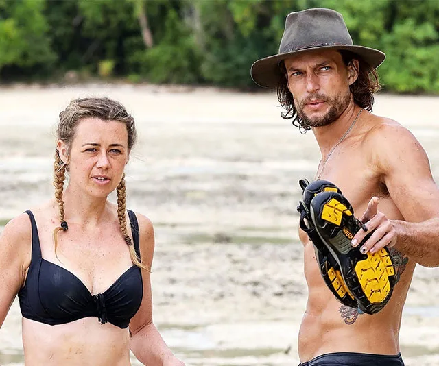 Two contestants in swimwear on Survivor 2020 set, one holding shoes, on a sandy beach with greenery in the background.