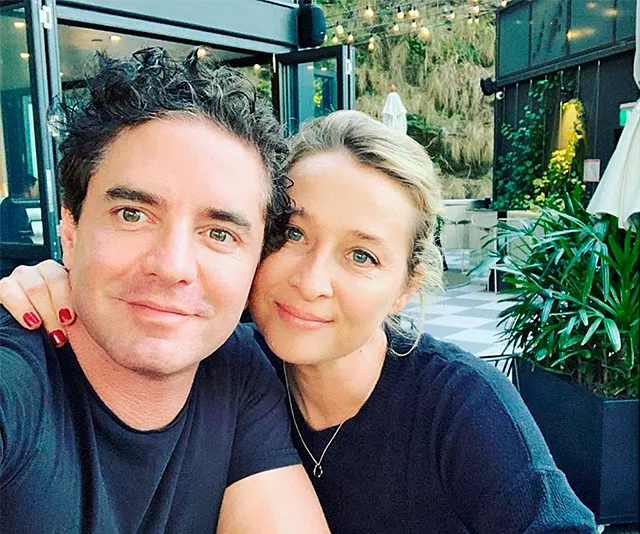A couple smiling at a well-lit outdoor café with greenery in the background.