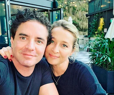 A couple smiling at a well-lit outdoor café with greenery in the background.