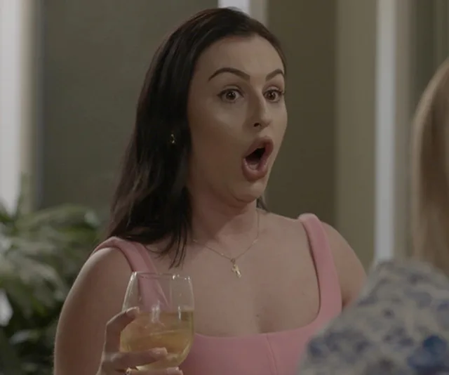 A woman in a pink dress looks surprised, holding a wine glass, indoors.