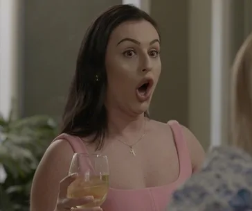 A woman in a pink dress looks surprised, holding a wine glass, indoors.