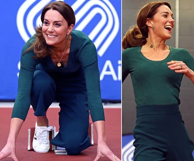 "Person in green outfit kneeling at a track start, smiling, with arms ready to run in an indoor athletics setting."