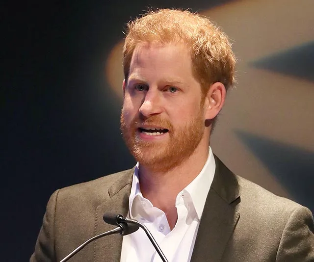 A man with red hair and a beard speaks at a podium during an event, wearing a suit and open-collared shirt.