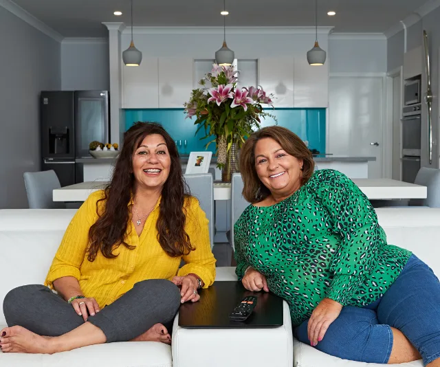 Two women sitting on a couch in a cozy living room with flowers in the background.
