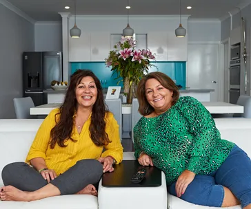 Two women sitting on a couch in a cozy living room with flowers in the background.