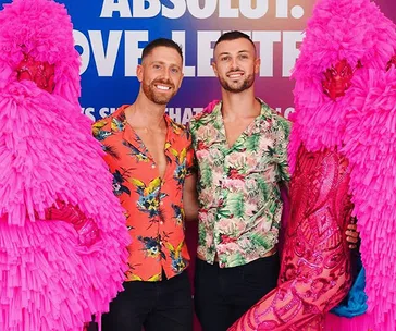 Two men in floral shirts pose with people in vibrant pink costumes at Sydney Mardi Gras 2020.