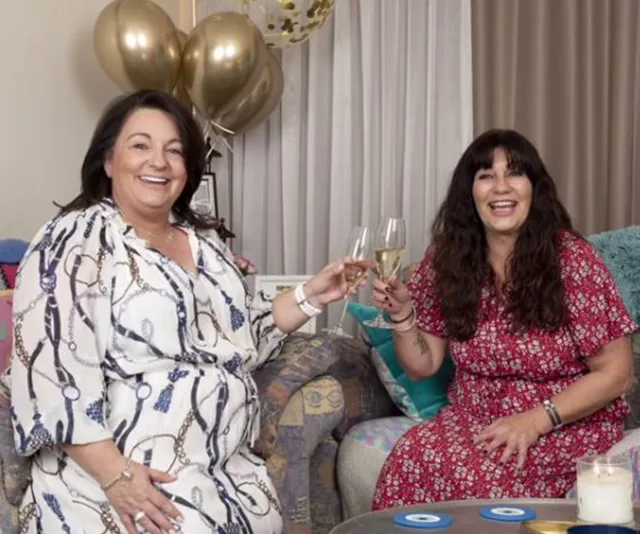Two women sitting on a couch, smiling, holding glasses of champagne, with golden balloons in the background.