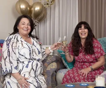 Two women sitting on a couch, smiling, holding glasses of champagne, with golden balloons in the background.