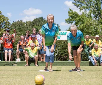 Women playing lawn bowls with an audience cheering under a bright, sunny sky.
