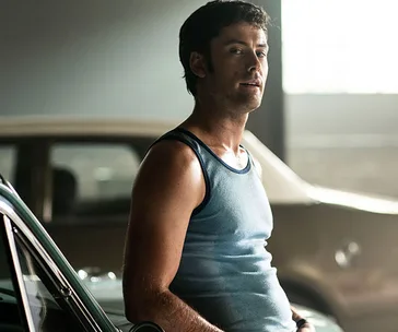 Man leaning against a car in a dimly lit garage, wearing a blue tank top.