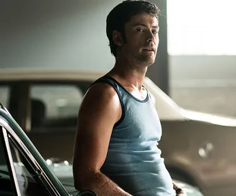 Man leaning against a car in a dimly lit garage, wearing a blue tank top.