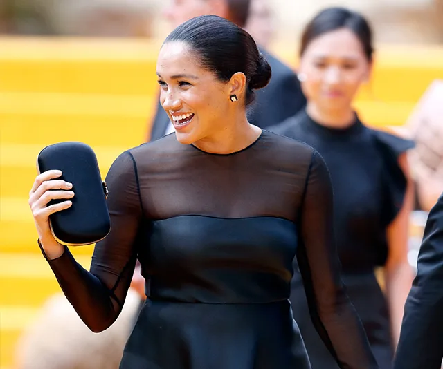 A person in a black evening gown smiles while holding a clutch, with people in the background, on a yellow-carpeted event.