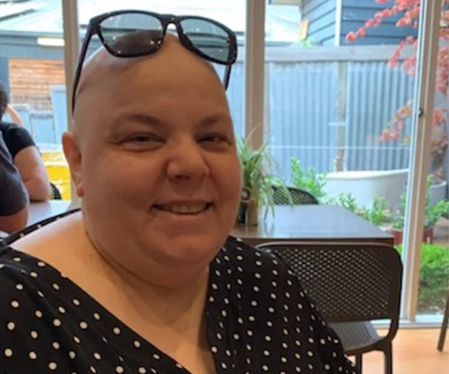 Smiling person sitting indoors, wearing sunglasses on head and polka dot top, with a shaved head, by a window.