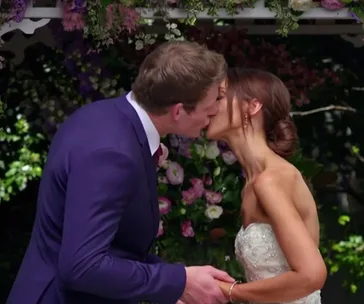 A bride and groom share a kiss at an outdoor wedding ceremony, surrounded by flowers and greenery.