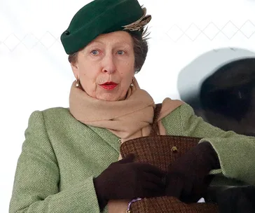 Woman in a green coat and hat with a beige scarf and gloves, sitting and holding a bag.