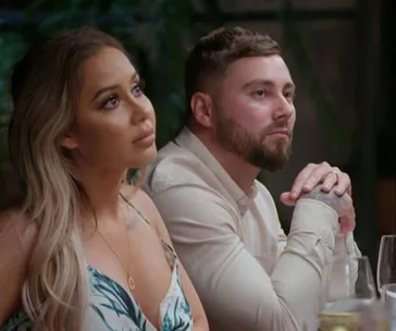 Two people sitting at a table, one with long hair in a patterned outfit, and the other with short hair wearing a light shirt.