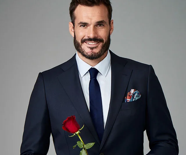 Man in a suit holding a red rose, smiling.