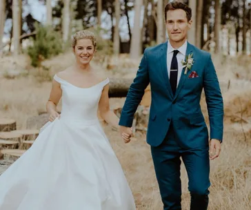 A bride in a white dress and groom in a blue suit holding hands outdoors in a rustic setting.