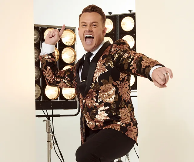 Man in a black and gold floral jacket posing energetically in front of stage lights.