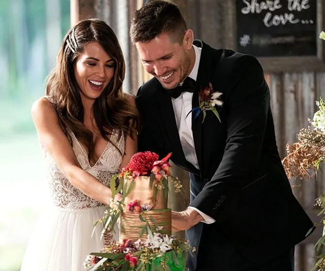 Bride in white dress and groom in tuxedo joyfully cutting a two-tiered floral wedding cake indoors.
