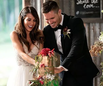 Bride in white dress and groom in tuxedo joyfully cutting a two-tiered floral wedding cake indoors.