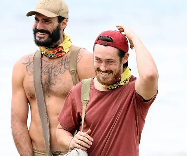 Two contestants from "Survivor 2020" stand on the beach smiling, one shirtless with tattoos, the other in a red shirt and cap.