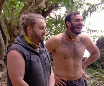 Two men outdoors in a tropical forest, one shirtless with tattoos, both wearing tribal-style bandanas.