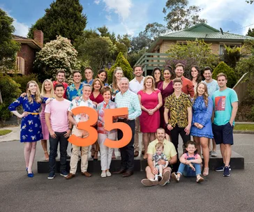 Group photo of "Neighbours" cast celebrating 35 years, with diverse members holding a large "35" in front of TV set houses.