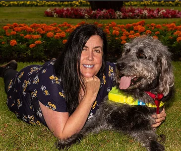 A woman lies on grass beside a fluffy gray dog with a guide harness, surrounded by colorful flowers.