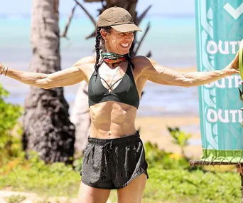 A woman in a swimsuit and cap stands outdoors on a tropical beach, smiling with arms raised next to a colorful banner.