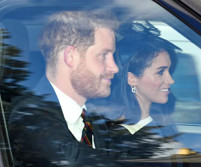 Couple in a car smiling, she wears a fascinator hat, both dressed formally.