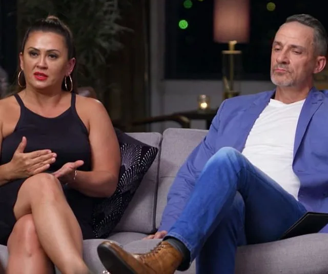 Couple sitting on a couch, woman in black dress gesturing, man in blue blazer listening.
