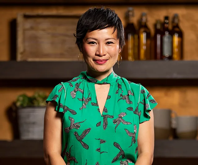 Poh, a contestant from MasterChef 2020, smiles in a green bird-patterned dress, standing in a kitchen setting.