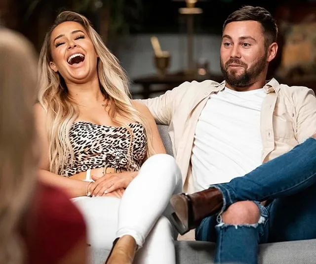 A couple sits on a couch, with the woman laughing and the man smiling, in a relaxed indoor setting.