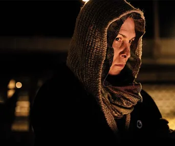 Person wearing a hooded sweater and scarf, gazing intently with a serious expression in a dimly lit setting.