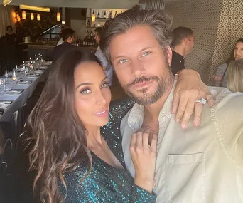 Couple posing together in a restaurant, smiling with warm lighting and other diners in the background.