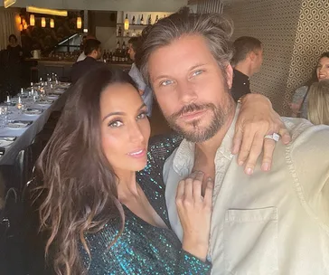 Couple posing together in a restaurant, smiling with warm lighting and other diners in the background.