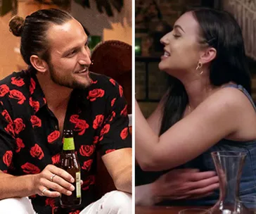 A man and woman are talking and smiling at a social event, each holding a drink in a lively setting.