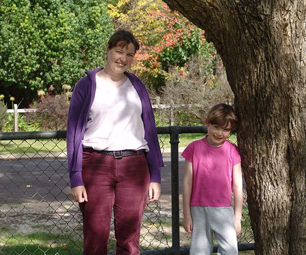 Two people, an adult and a child, stand beside a tree smiling, with autumn foliage in the background.