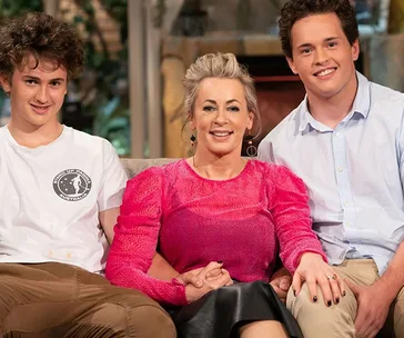 A woman in a pink top sits on a couch with two young men, one in a white t-shirt and the other in a blue shirt.