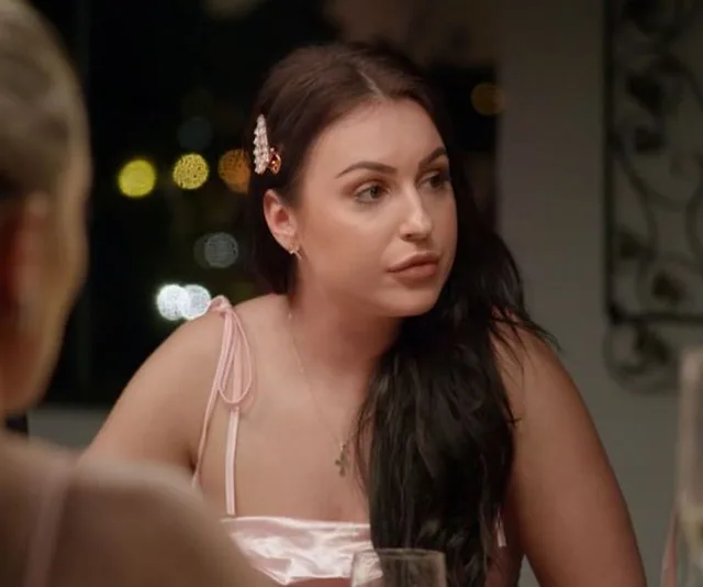 A woman with dark hair sits at a table, wearing a pink dress and a pearl hair clip, looking to the side.