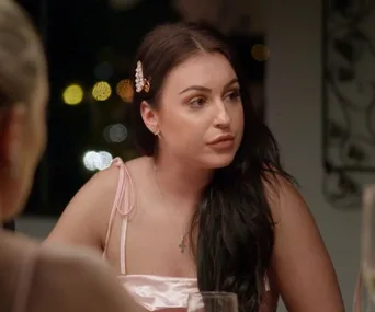 A woman with dark hair sits at a table, wearing a pink dress and a pearl hair clip, looking to the side.
