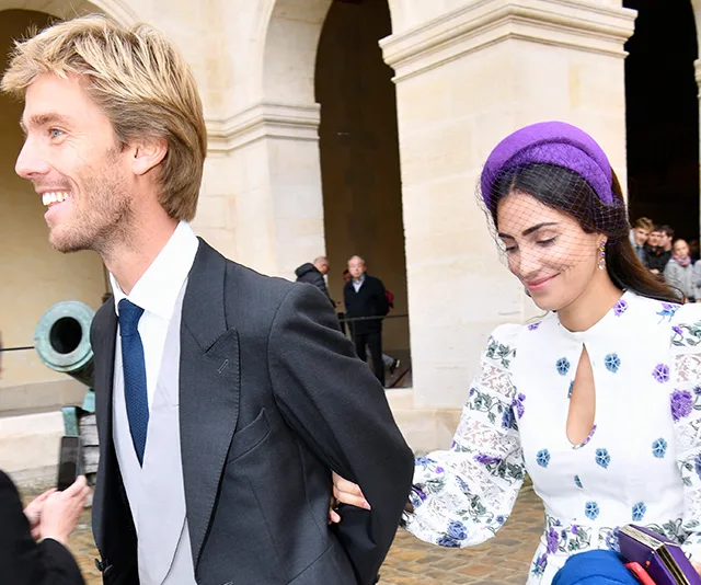 Couple walking arm-in-arm, man in a suit, woman in floral dress and purple hat, at an outdoor event or ceremony.