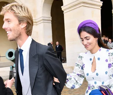 Couple walking arm-in-arm, man in a suit, woman in floral dress and purple hat, at an outdoor event or ceremony.