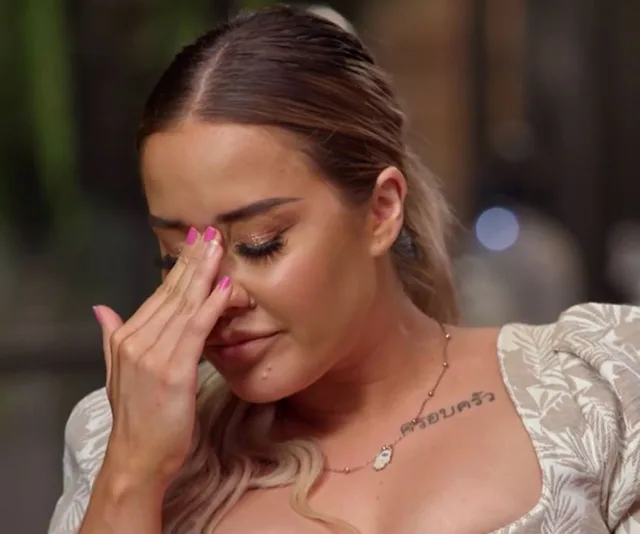 Woman with a tattoo on her shoulder, wiping tears from her eyes, dressed in a patterned off-shoulder top.