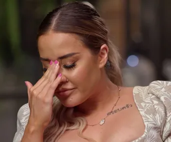 Woman with a tattoo on her shoulder, wiping tears from her eyes, dressed in a patterned off-shoulder top.